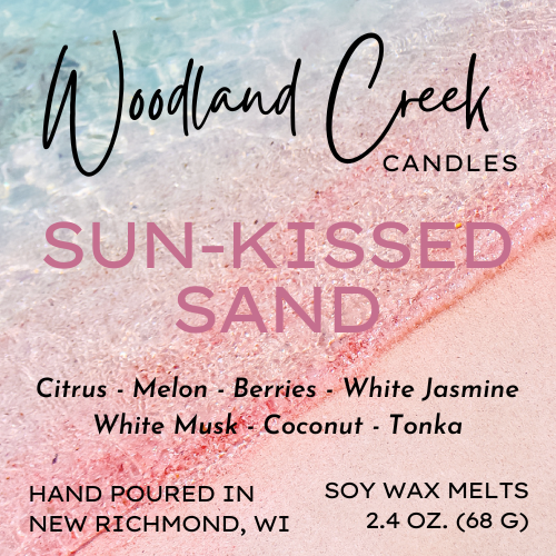 Product label for Woodland Creek Candles in the scent 'Sun-Kissed Sand'. The label features a textured background resembling a beach shoreline with pastel blue, pink, and beige tones.